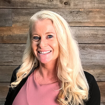 A woman with long blonde hair wearing a pink top and black jacket smiles in front of a wooden plank wall at Thrive Senior Living.