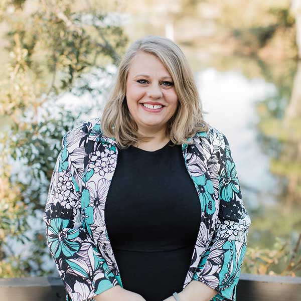 A woman with blonde hair wearing a black top and a floral jacket smiles outdoors with trees and water in the background, embodying the vibrant spirit of Thrive Senior Living.