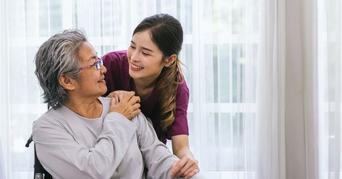 A young caregiver in purple scrubs from Thrive Senior Living smiles and gently supports an older woman in glasses sitting in a wheelchair indoors.