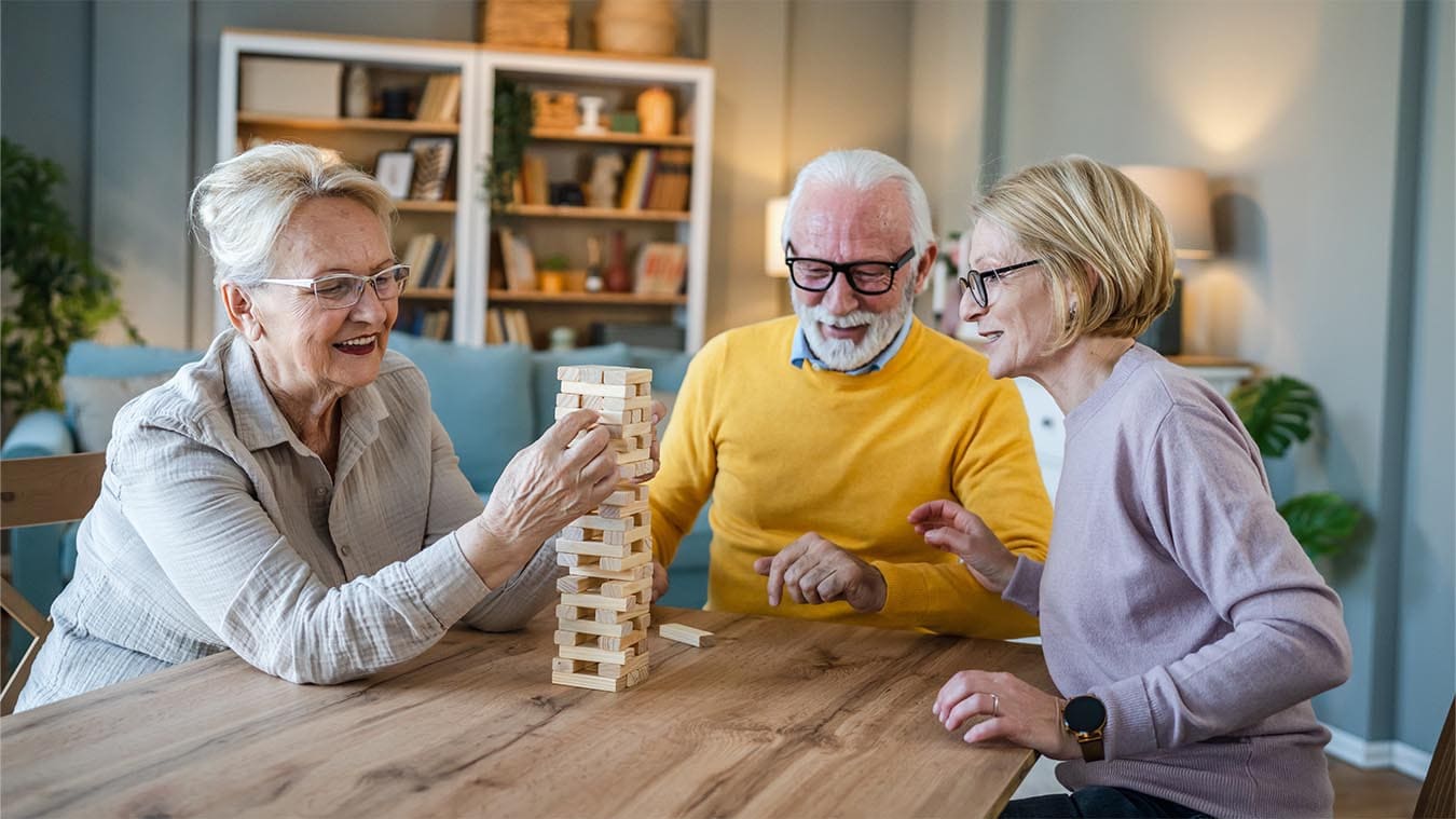Three older adults sit at a wooden table, smiling and playing a game of Jenga in a cozy, well-lit Thrive Senior Living community.
