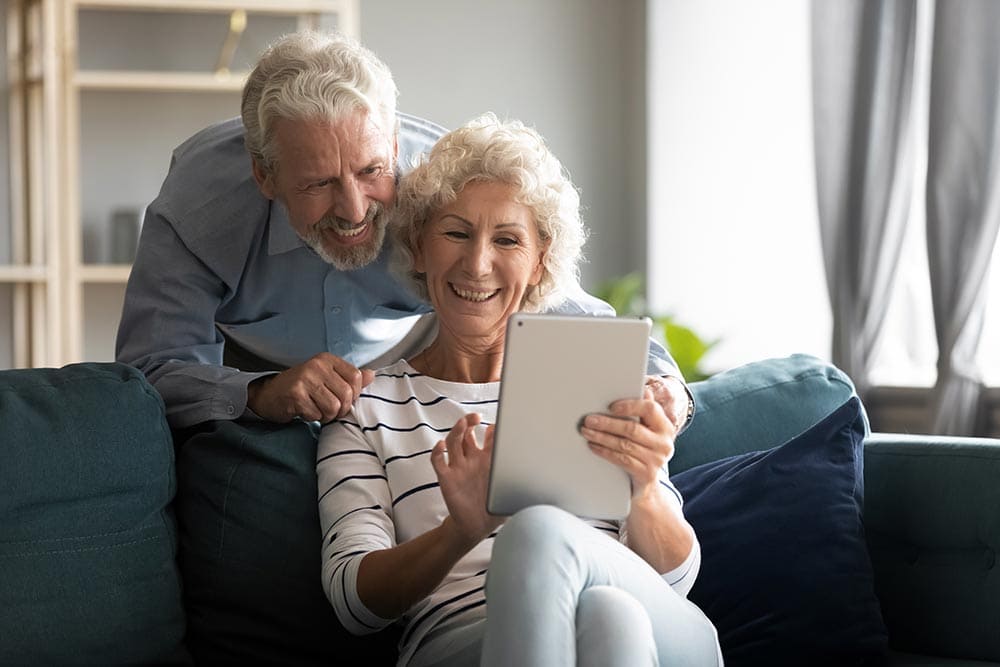 An older couple sits on a couch at Thrive Senior Living; the woman holds a tablet and smiles while the man leans over her shoulder, both looking at the screen.