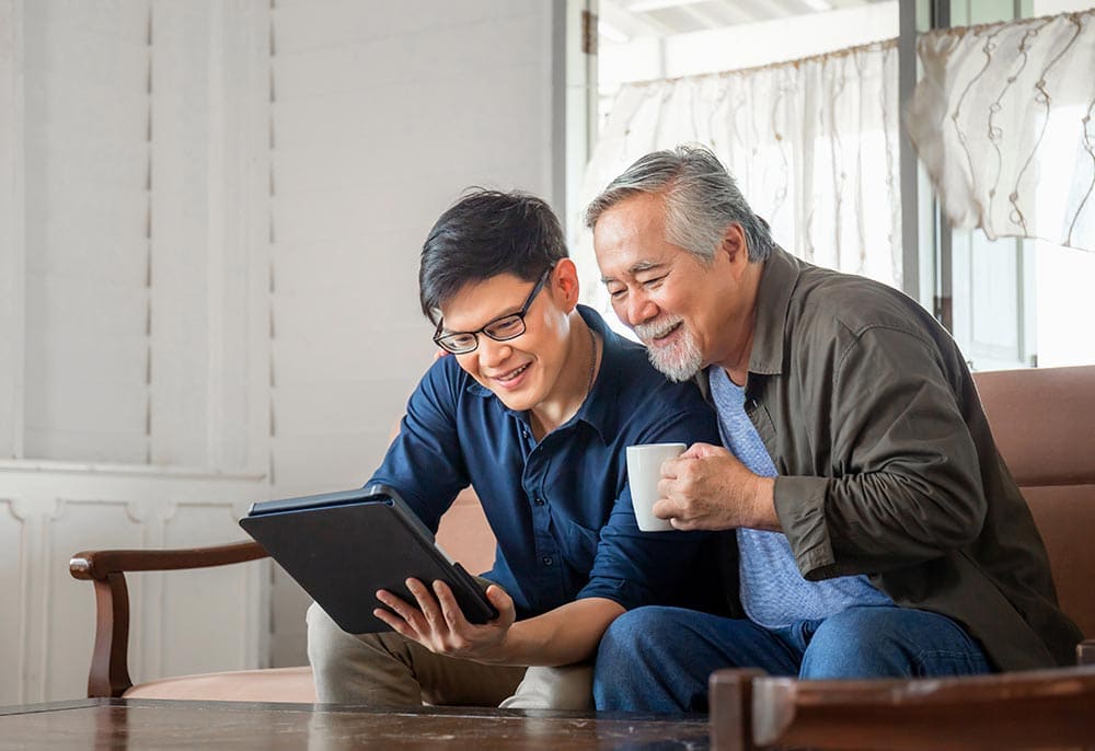 An older man and a younger man sit on a couch, smiling as they look at a tablet together at Thrive Senior Living; the older man holds a mug.