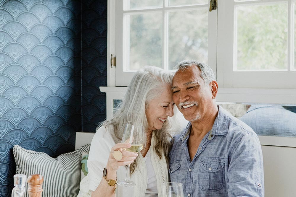 An older couple sits closely together, smiling and laughing, as one holds a glass of white wine in a bright, cozy room with blue patterned walls—reflecting the warm atmosphere found at Thrive Senior Living.