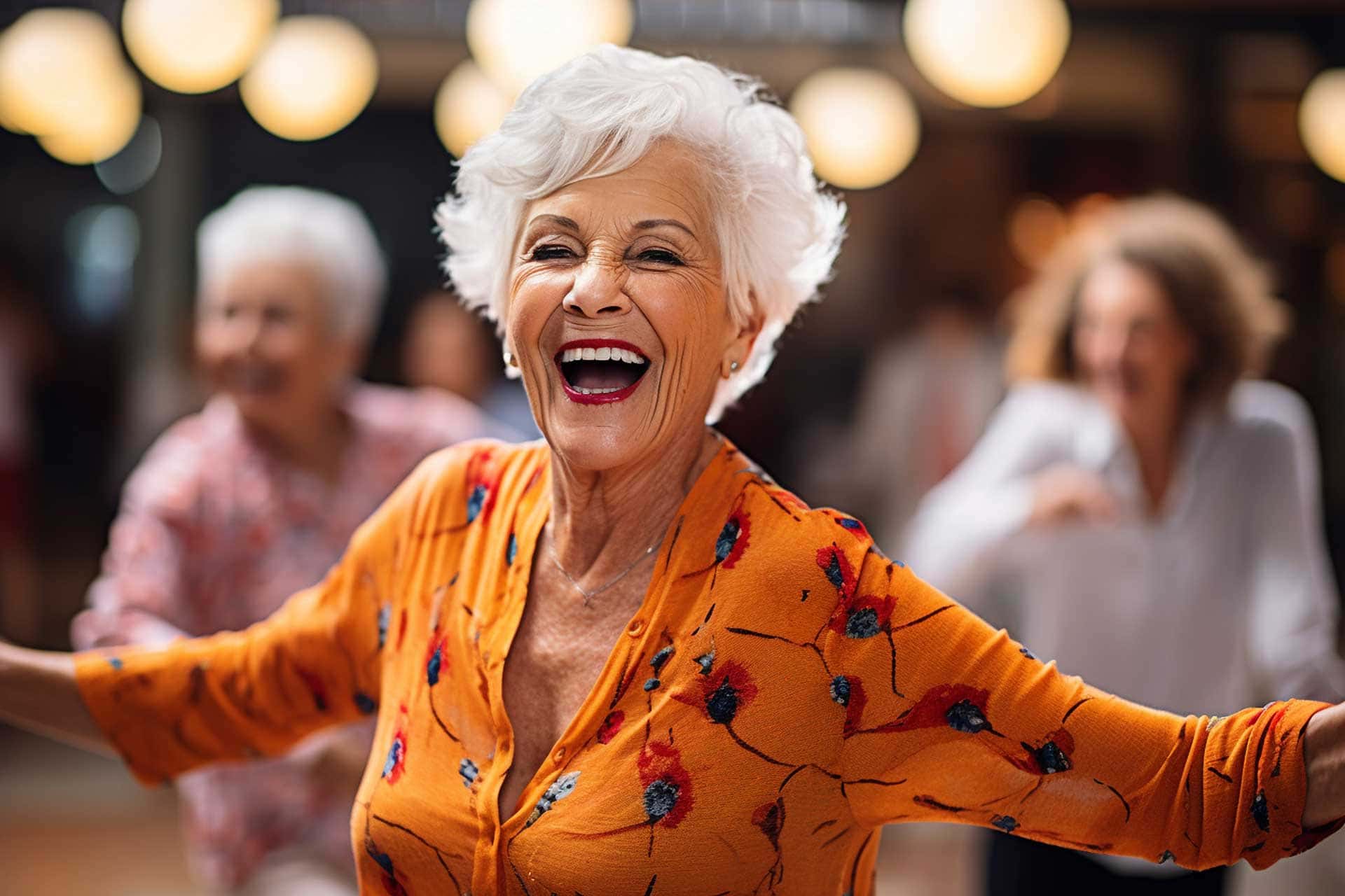 Older woman with short white hair, wearing an orange floral blouse, smiling and dancing among a group of people indoors at Thrive Senior Living.