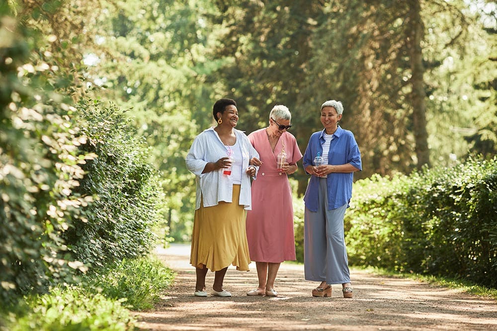 Three older women walk together on a sunlit path in a park, holding drinks and chatting, surrounded by greenery and trees—a perfect moment of connection inspired by Thrive Senior Living.