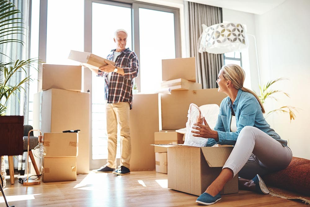 Two people in a bright room unpacking moving boxes at Thrive Senior Living, with several boxes scattered around and sunlight streaming through large windows.