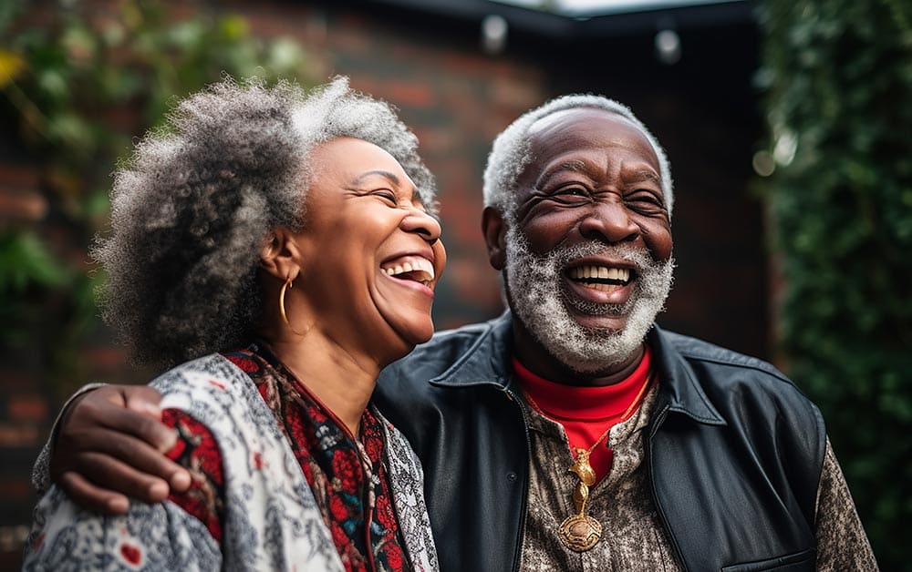 An older couple stands outdoors, smiling and laughing together. The background is blurred with greenery and a brick wall visible, capturing the joyful spirit found at Thrive Senior Living.