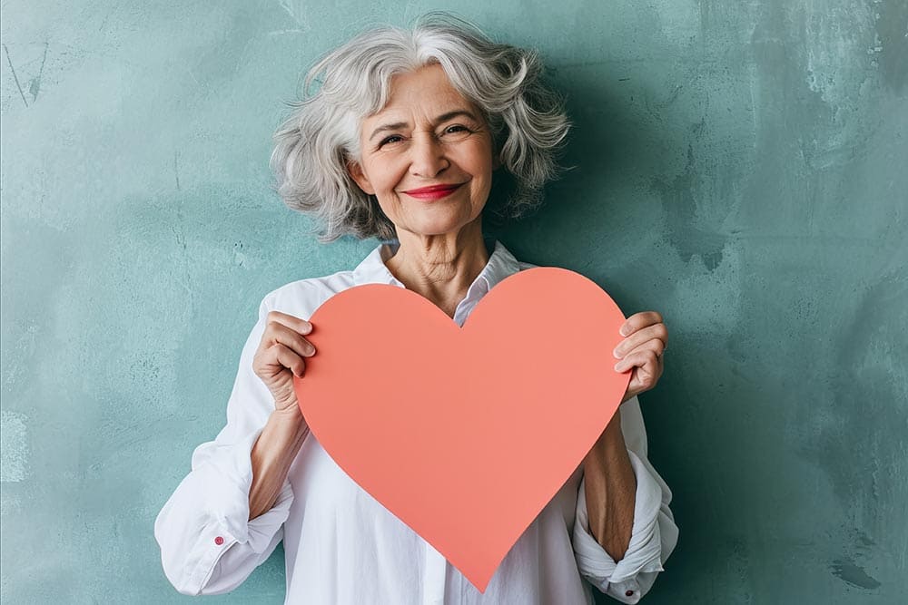 Older woman with gray hair, wearing a white shirt, smiling and holding a large red paper heart in front of a textured green background—reflecting the joyful spirit found at Thrive Senior Living.