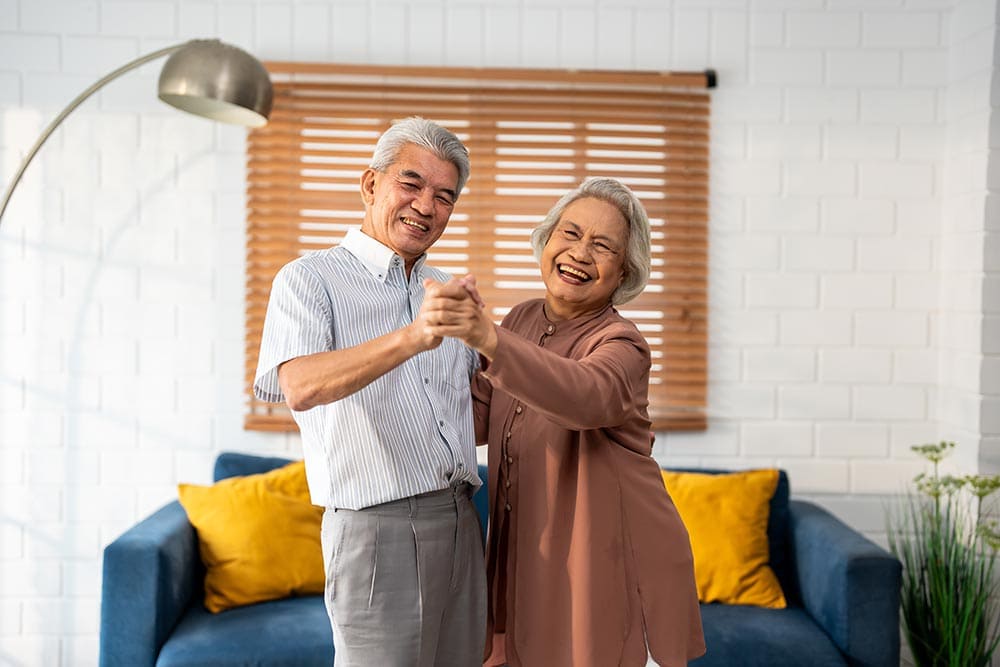 Two older adults smile and hold hands while dancing in a cozy living room with a blue couch and yellow pillows—an uplifting moment that captures the spirit of Thrive Senior Living.