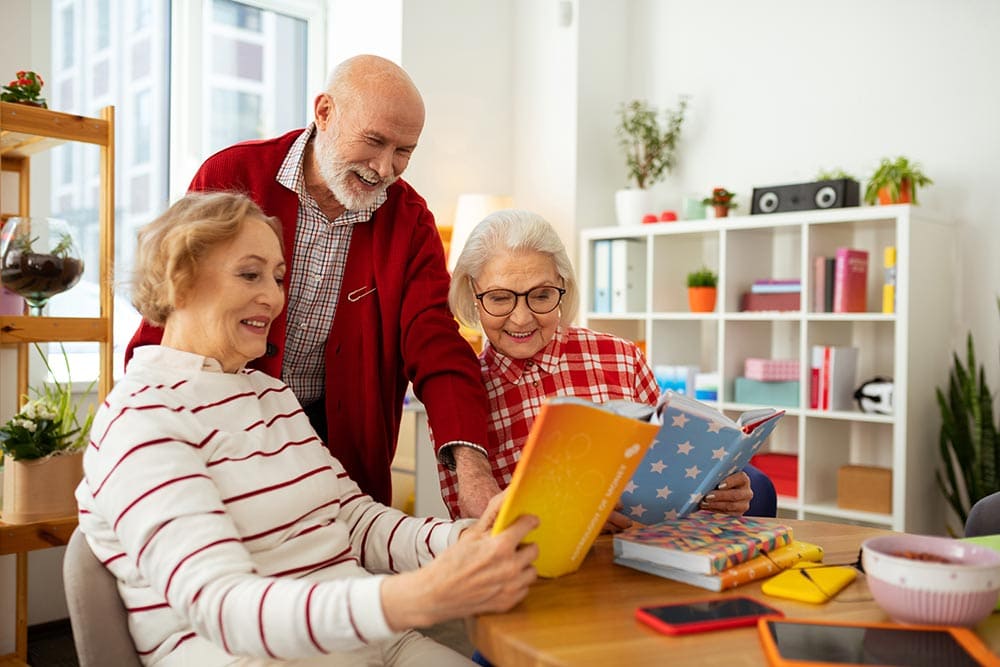 Three older adults sit at a table in a bright room at Thrive Senior Living, smiling and looking at colorful books together.