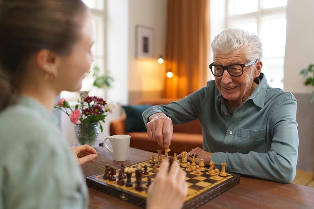 An older man and a younger woman play chess at a wooden table in a bright Thrive Senior Living room, with flowers, a mug, and large windows in the background.
