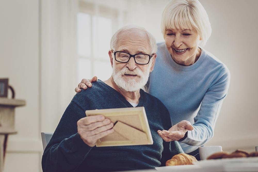 An older man with glasses looks at a framed photo while an older woman stands beside him, smiling and placing a hand on his shoulder, enjoying a warm moment together at Thrive Senior Living.