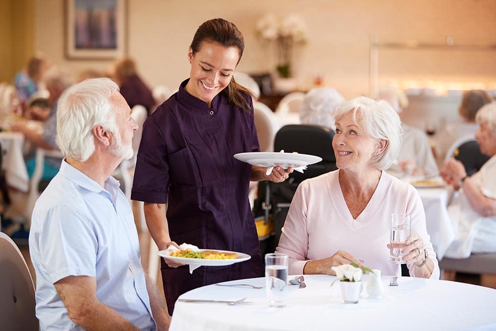 A server hands plates of food to an elderly man and woman sitting together at a dining table in a Thrive Senior Living communal dining area.