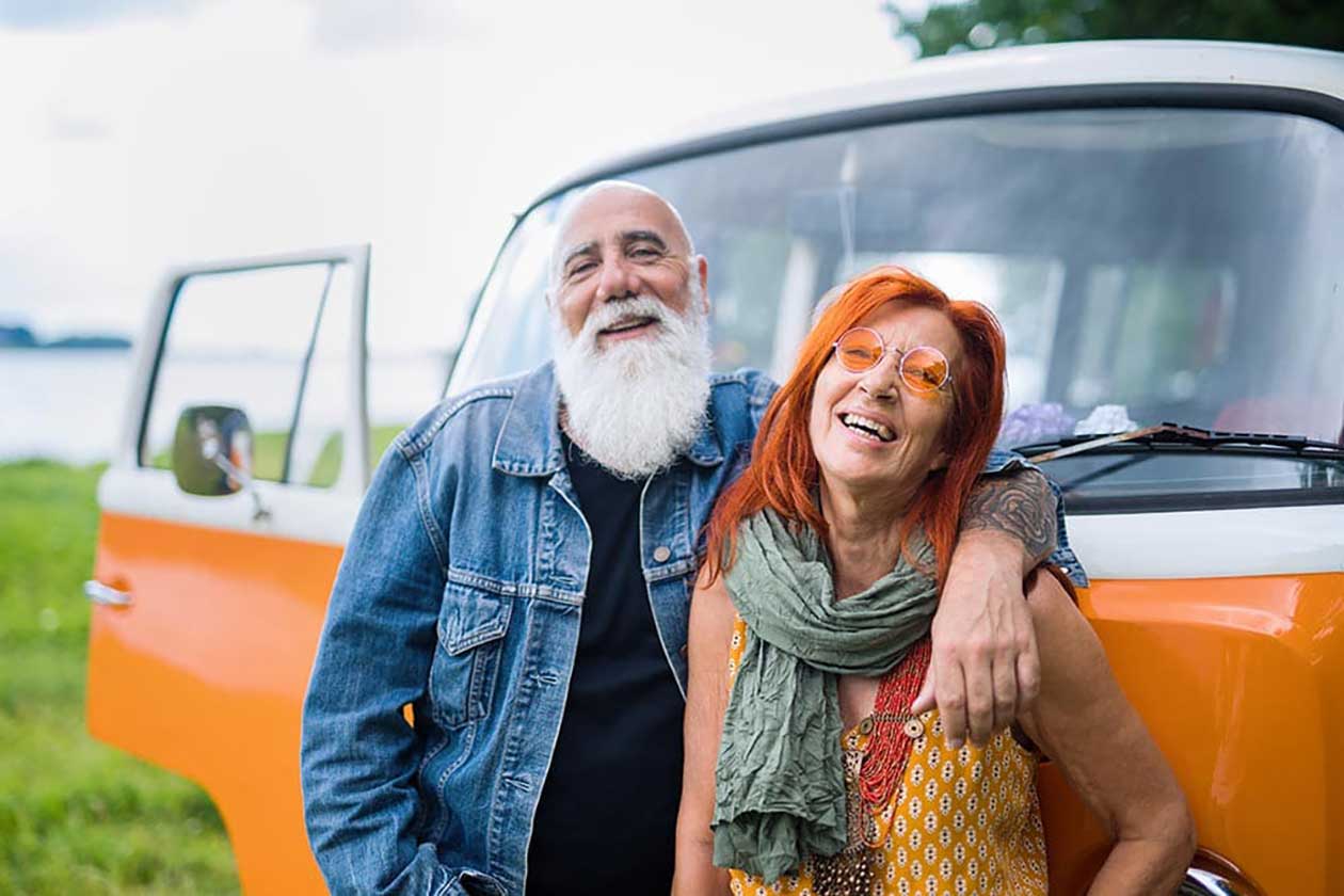 An older man and woman stand smiling beside an orange van, with the man's arm around the woman's shoulders. The open van door and lush greenery capture a joyful moment, reflecting the vibrant lifestyle at Thrive Senior Living.