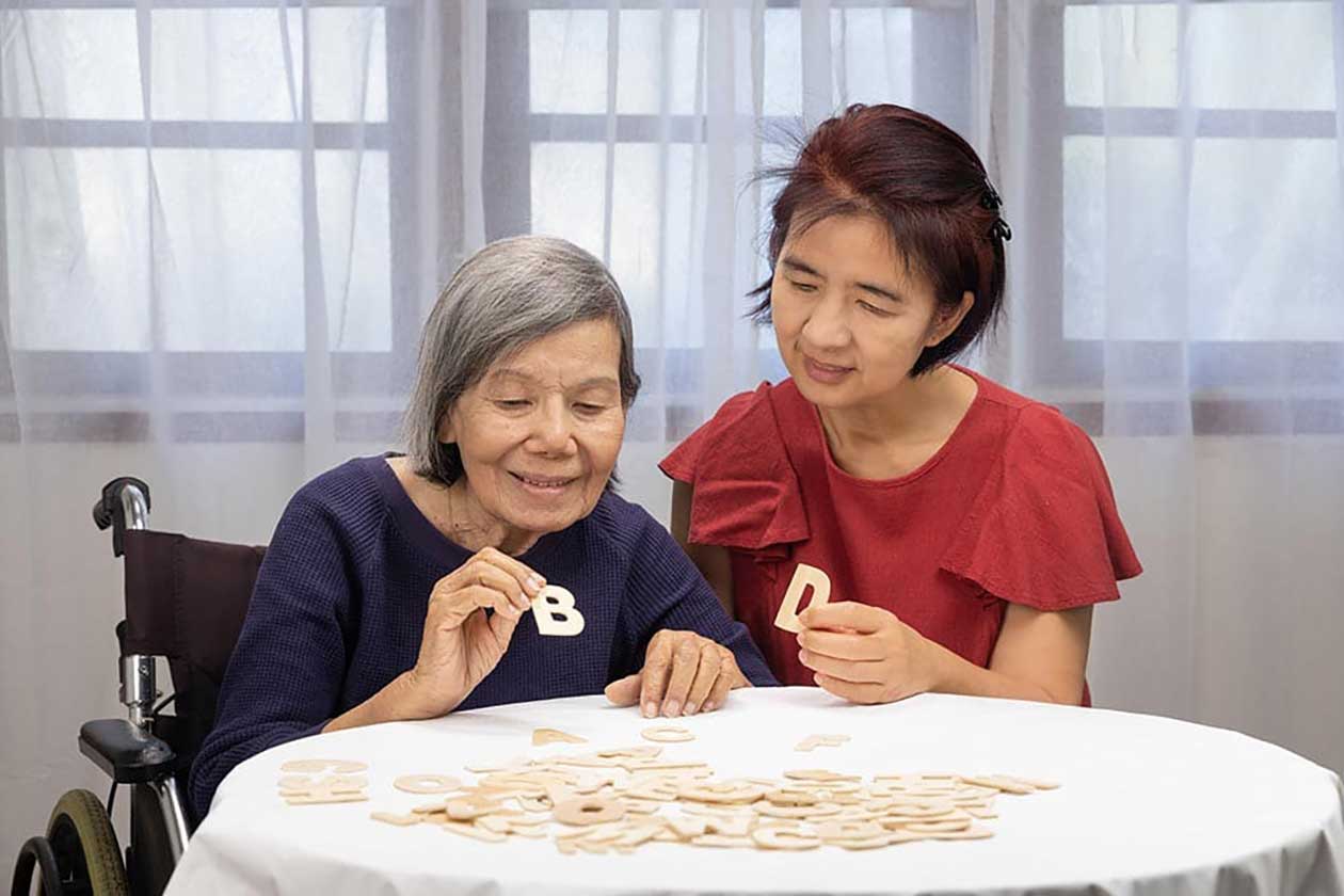 Two women sit at a table covered with wooden letters at Thrive Senior Living; one holds a letter "B" while the other displays a letter "D.