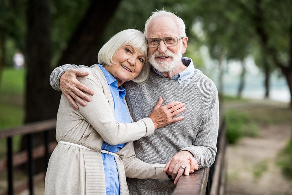 An elderly couple stands on a wooden bridge outdoors, smiling and embracing each other amid trees and greenery—a warm moment that reflects the joyful spirit of Thrive Senior Living.