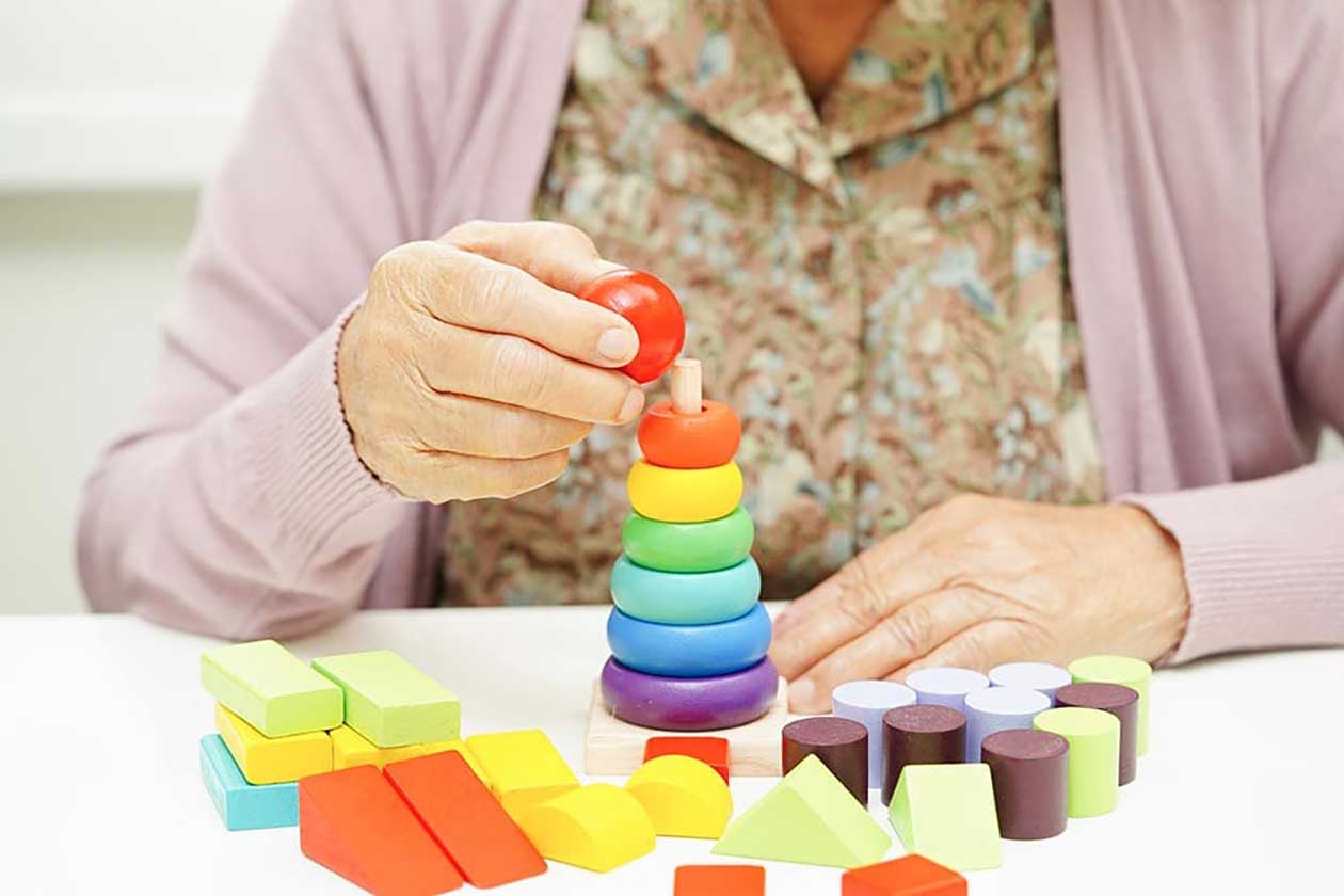 An elderly person at Thrive Senior Living arranges colorful wooden stacking rings and geometric blocks on a white table.