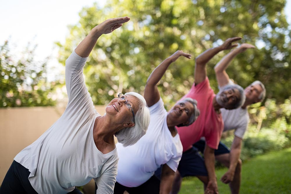 A group of older adults at Thrive Senior Living doing side stretch exercises outdoors in a line, with trees and greenery in the background.