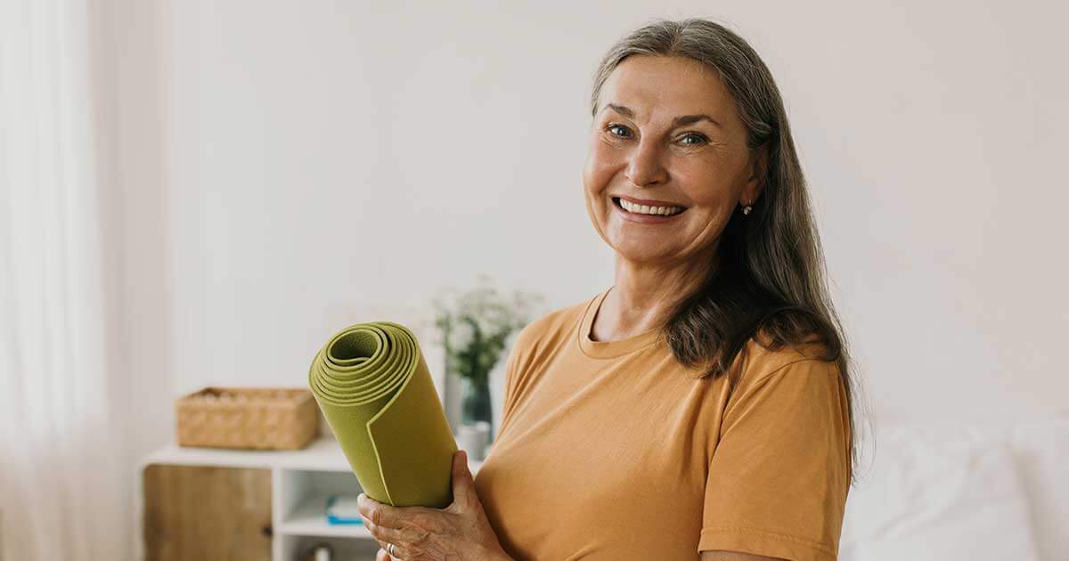 A woman with long gray hair, wearing an orange shirt, smiles while holding a rolled-up green yoga mat indoors at Thrive Senior Living.