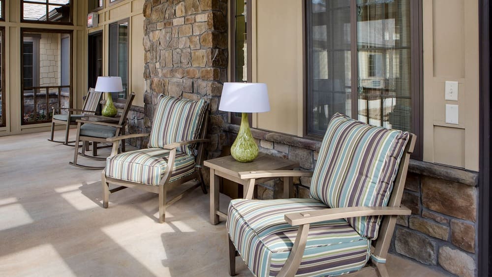 Outdoor patio at Thrive Senior Living features striped cushioned chairs, small side tables, and green table lamps, set against a stone and beige wall.