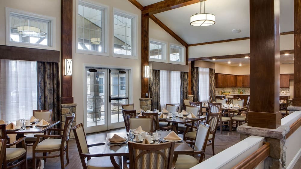 A bright dining room at Thrive Senior Living, with neatly arranged tables and chairs set with plates, glasses, and napkins, featuring large windows, wood accents, and modern light fixtures.