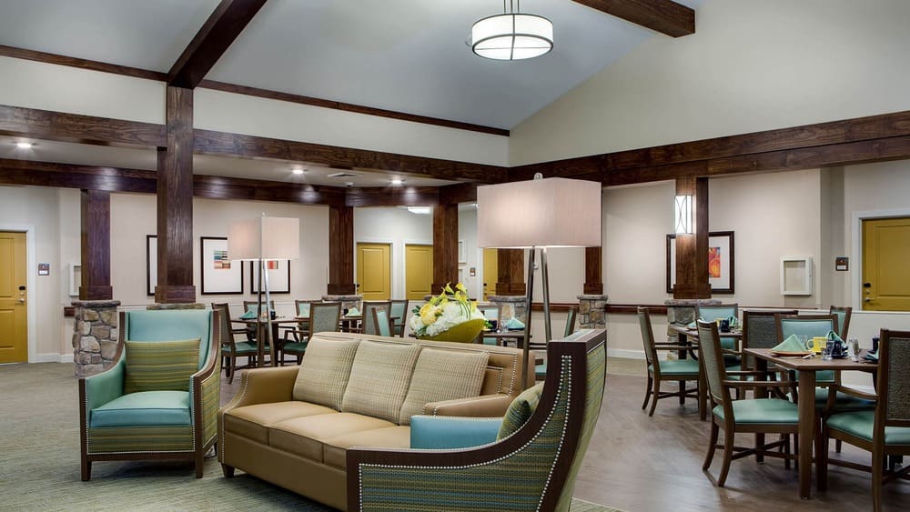 Spacious lounge and dining area at Thrive Senior Living, featuring upholstered chairs, a sofa, tables set for dining, and soft lighting under a high ceiling with wooden beams.