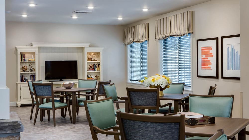 A modern communal room at Thrive Senior Living with tables and chairs, a flat-screen TV on a white entertainment center, bookshelves, two windows with blinds, and framed artwork on the wall.