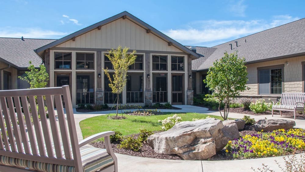 A landscaped courtyard with benches, trees, flowers, and large rocks welcomes residents at Thrive Senior Living, surrounded by a single-story building with large windows and a peaked roof.