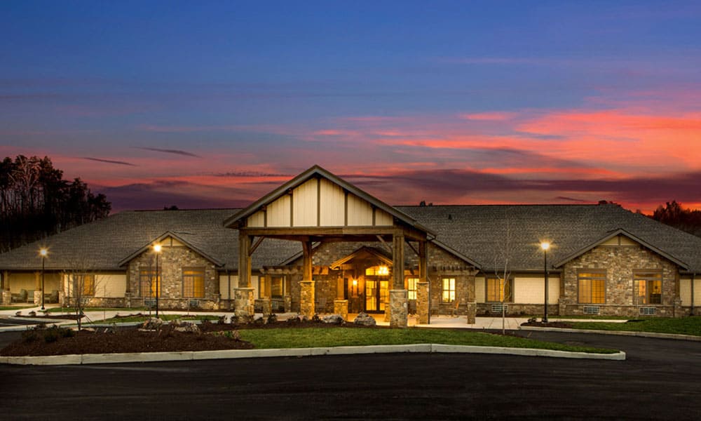 A single-story Thrive Senior Living building with stone and wood accents is illuminated at dusk, set against a colorful sunset sky, with a paved parking area in front.