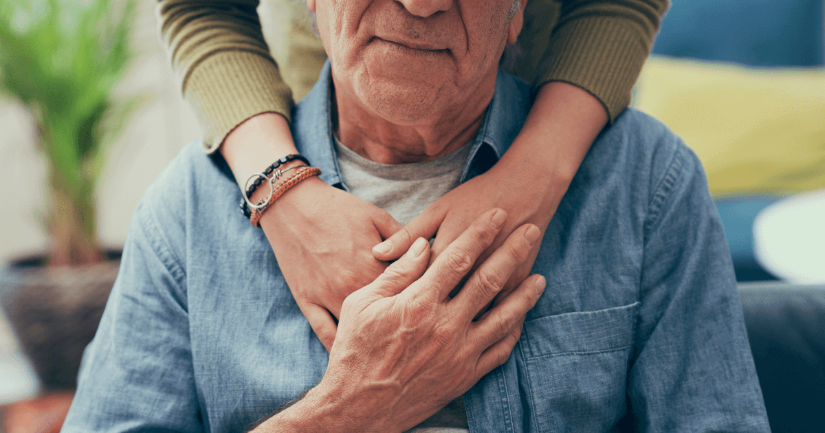 Older man with another's arms around him and he is touching the their hands.