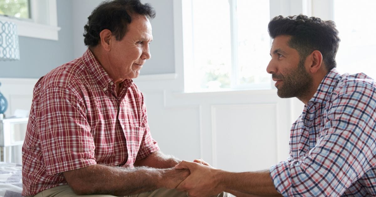 Two men sit facing each other indoors, holding hands and making eye contact, suggesting a serious or supportive conversation at Thrive Senior Living.