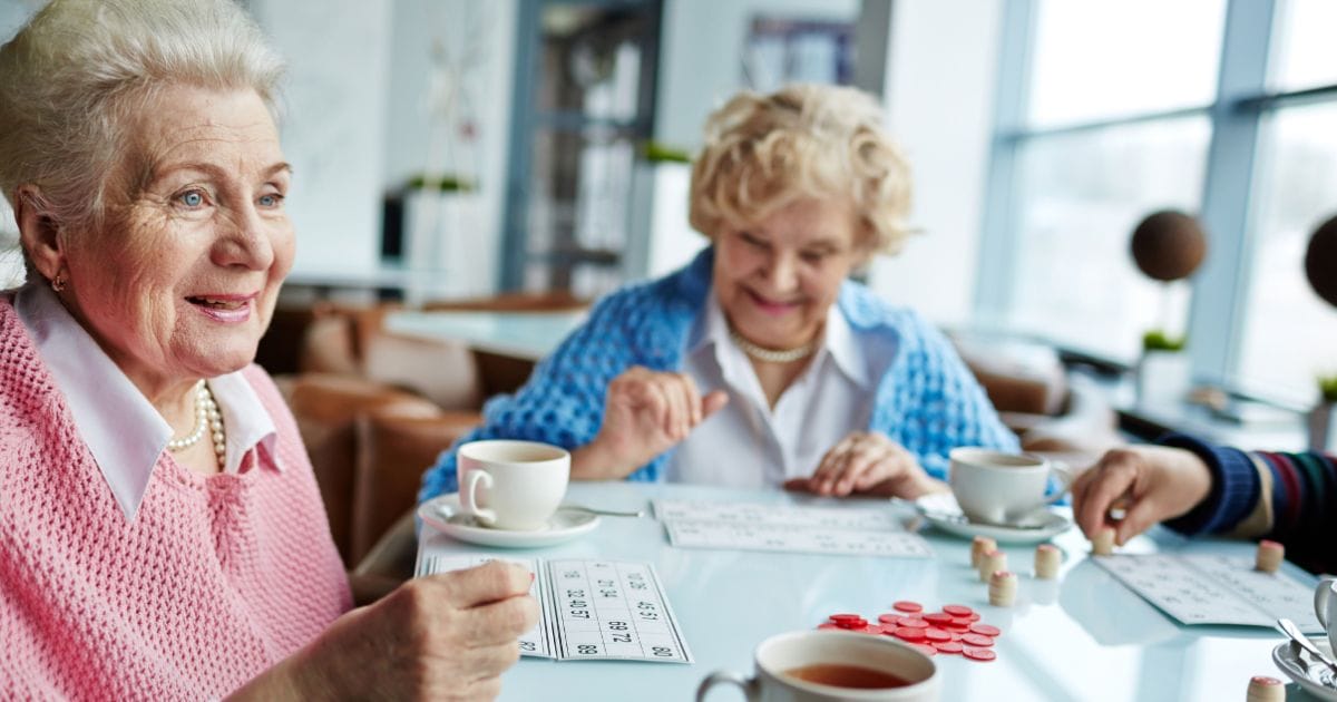 Two elderly women sit at a table with bingo cards, red chips, and teacups, smiling and engaged in a bright Thrive Senior Living community social setting.