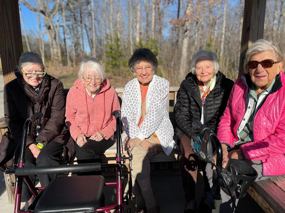 Five elderly women sit together on a wooden bench outdoors on a sunny day at Thrive Senior Living, with walkers positioned in front of them and bare trees visible in the background.