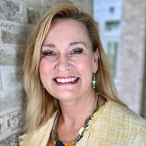 A woman with long blonde hair smiles at the camera, standing next to a light brick wall at Thrive Senior Living, wearing a yellow top with green earrings and a necklace.