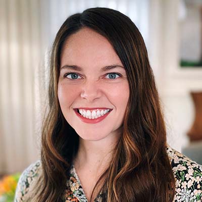 A woman with long brown hair and a floral top smiles at the camera in a softly lit indoor space at Thrive Senior Living, with elegant curtains behind her.