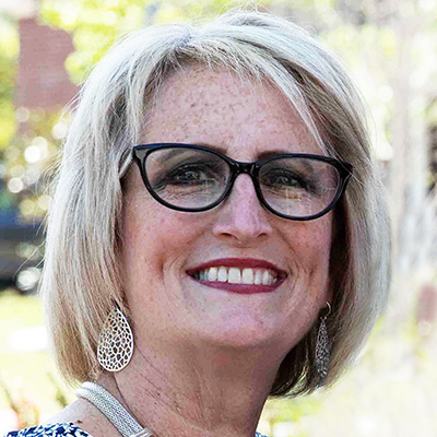 A woman with short blond hair, glasses, and dangling earrings smiles outdoors at Thrive Senior Living.