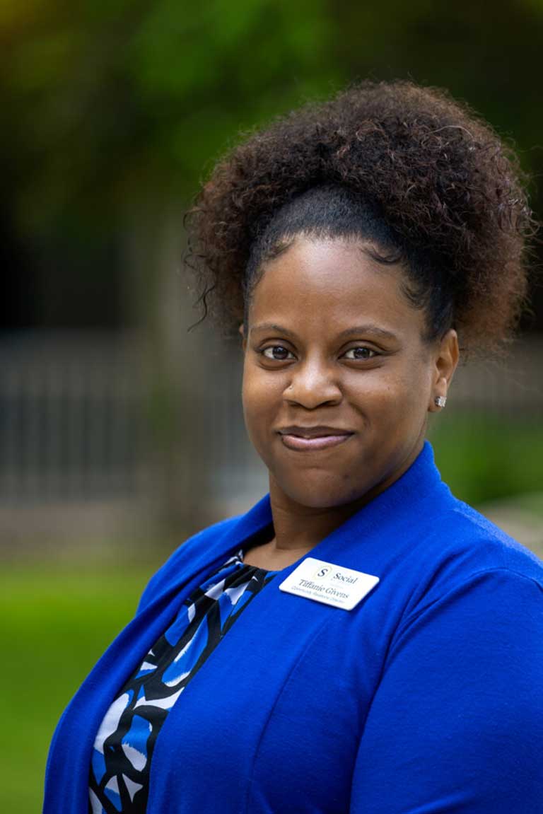 Woman wearing a blue blazer and patterned top, smiling at the camera with a Thrive Senior Living name tag on her chest and a blurred outdoor background.